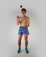 Man wearing blue shorts with red peach pattern, juggling peach in studio