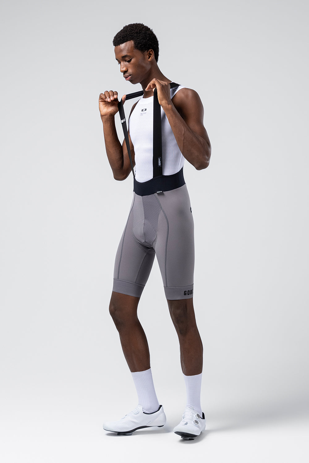 Male model adjusting Gobik bib short suspenders, wearing gray Kettle cycling shorts, side view