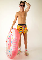 Man wearing yellow ChicknLegs shorts with colorful donut pattern, holding inflatable donut ring