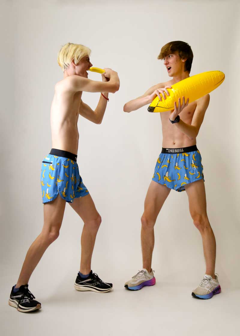 Two male models wearing ChicknLegs banana-print blue split running shorts playfully dueling with inflatable bananas