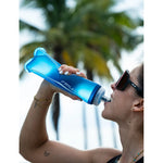 Woman drinking from Fitletic Flow Max Soft Flask 500ml blue bottle outdoors near palm trees.