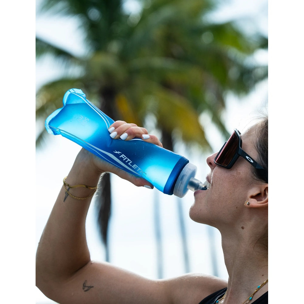 Woman drinking from Fitletic Flow Max Soft Flask 500ml blue bottle outdoors near palm trees.